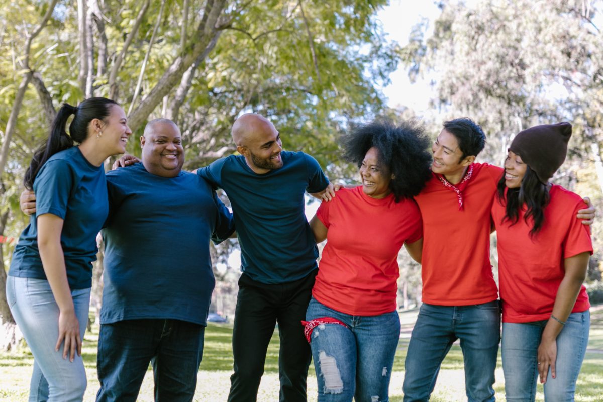 A photo of a group of six smiling middle-aged people, in a line, with arms around each other's waists. They appear to be in a park, wearing athletic clothing.