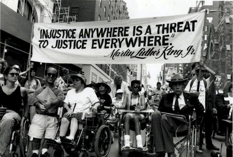 A black and white photo of people in wheelchairs holding a banner quoting Martin Luther King J. "Injustice anywhere is a threat to justice everywhere."